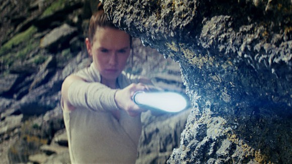 Actress Daisy Ridley as Star Wars character Rey holding a light blue lightsaber.