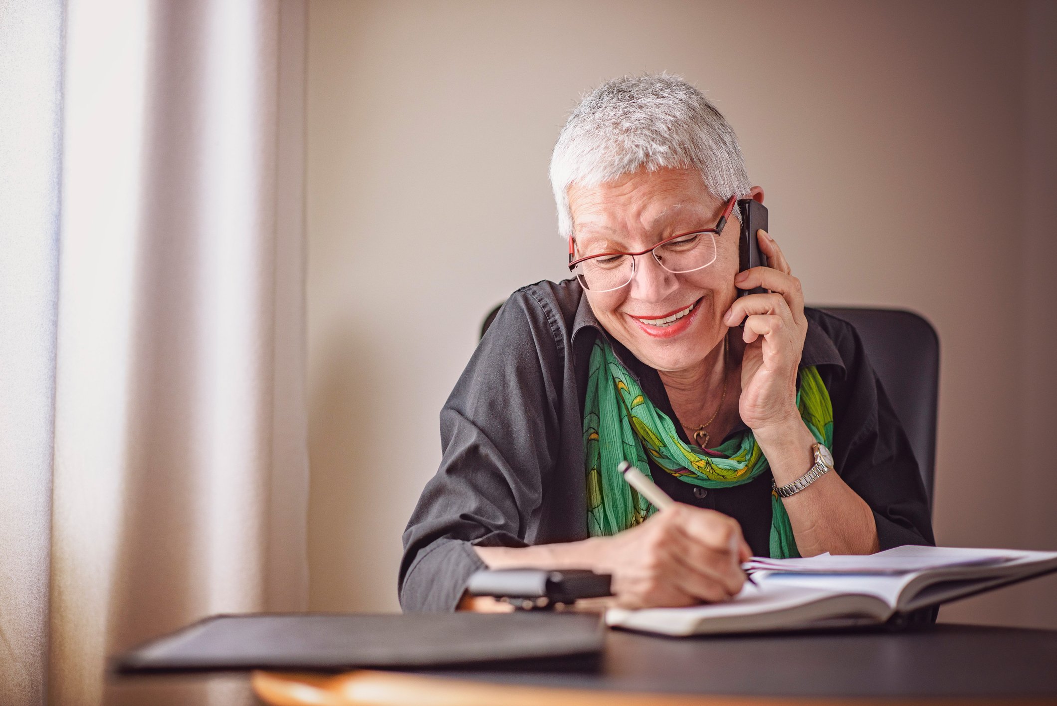 Happy senior old business woman writing down orders, talking on her cell phone