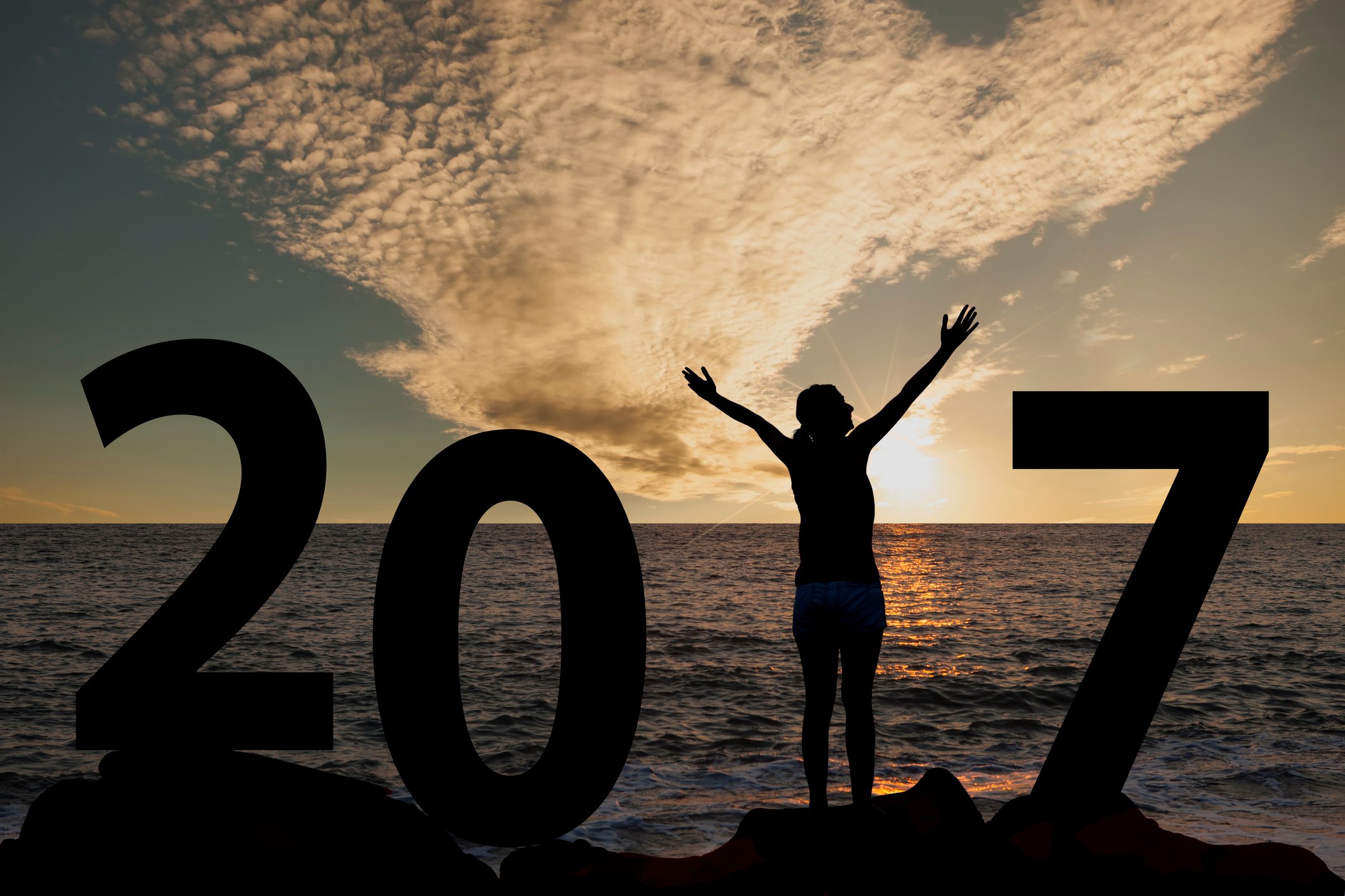 Image of a lady enjoying an ocean sunset with her arms held up signifying the number 1 in a large 2017 placed on a rocky shore. 