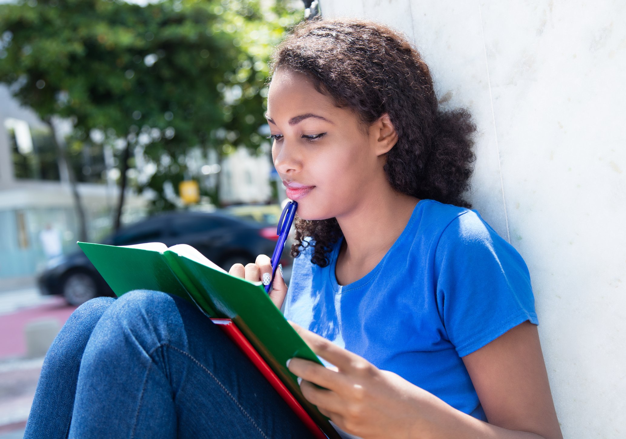 A young woman leans outdoors against a wall, knees drawn up, reading a book as she rests a pen against her chin.