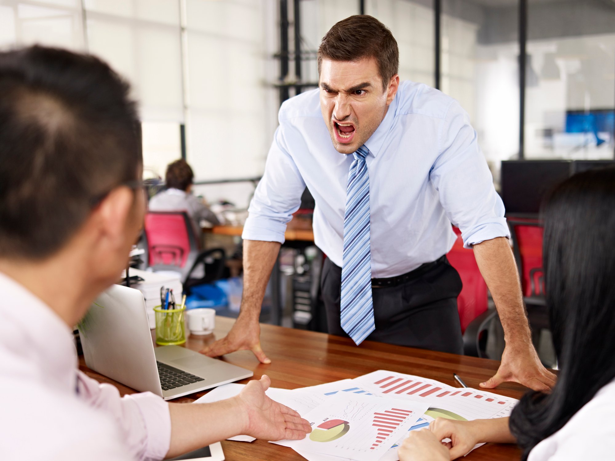 a young man in a tie yells at someone across a desk, who points to a company document, arguing.