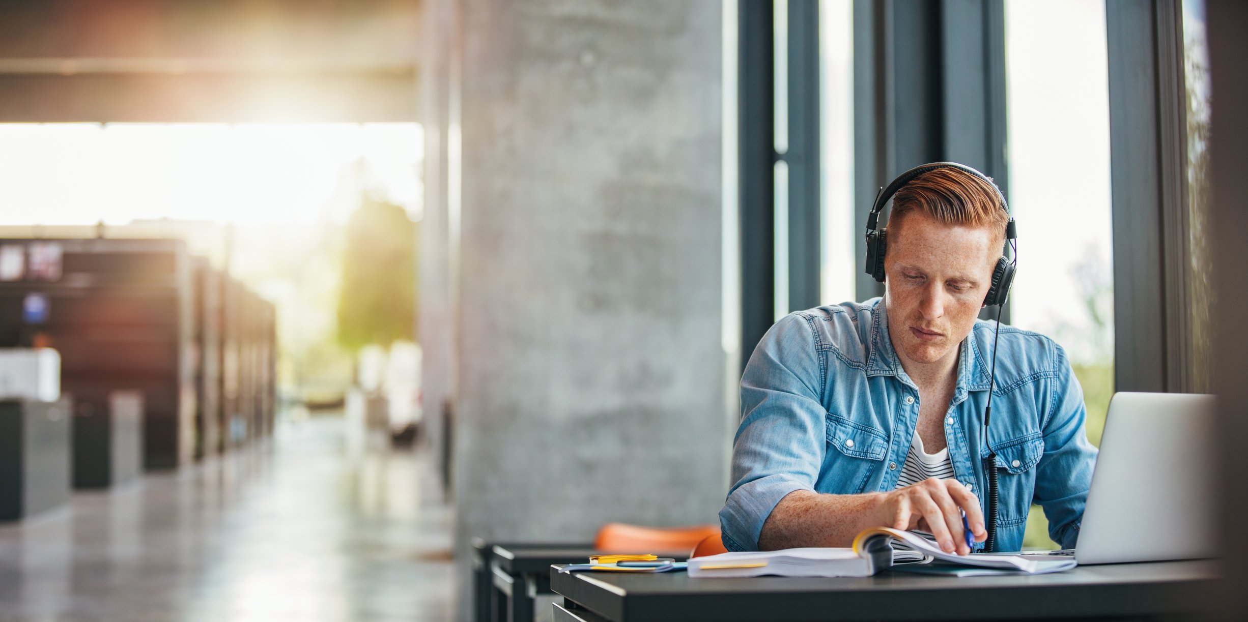 A young man with headphone on sits at a desk working on a laptop.