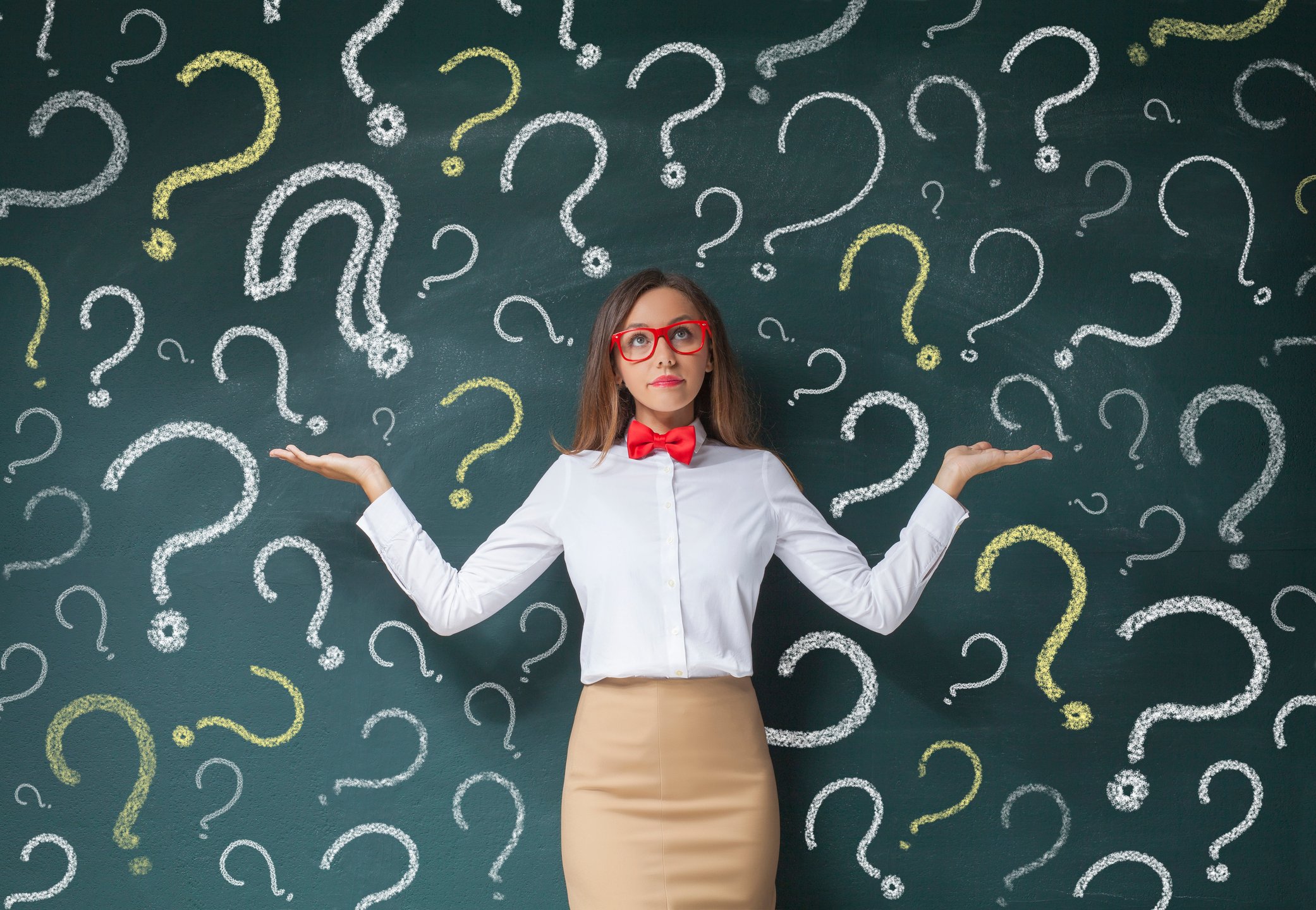 A businesswoman standing in front of a chalkboard with question marks drawn on it while she holds her hands up to the side.