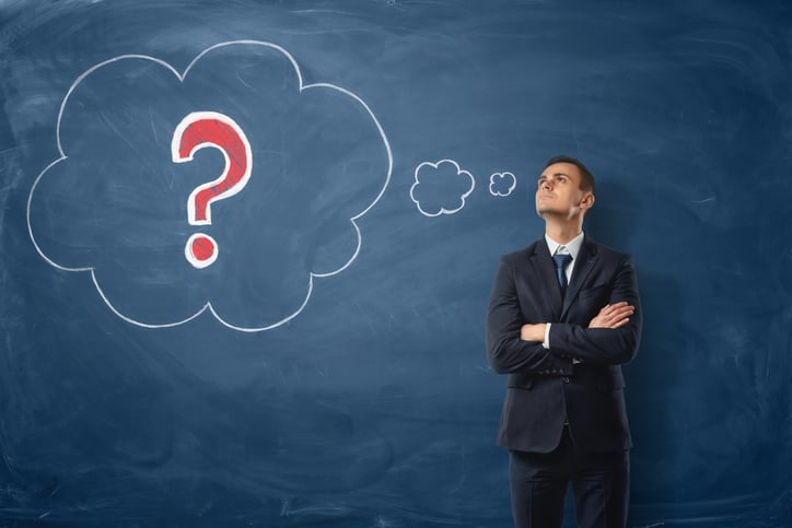 A businessman standing in front of a chalkboard with his arms crosses thinking as a thought bubble with a question mark is drawn behind him.