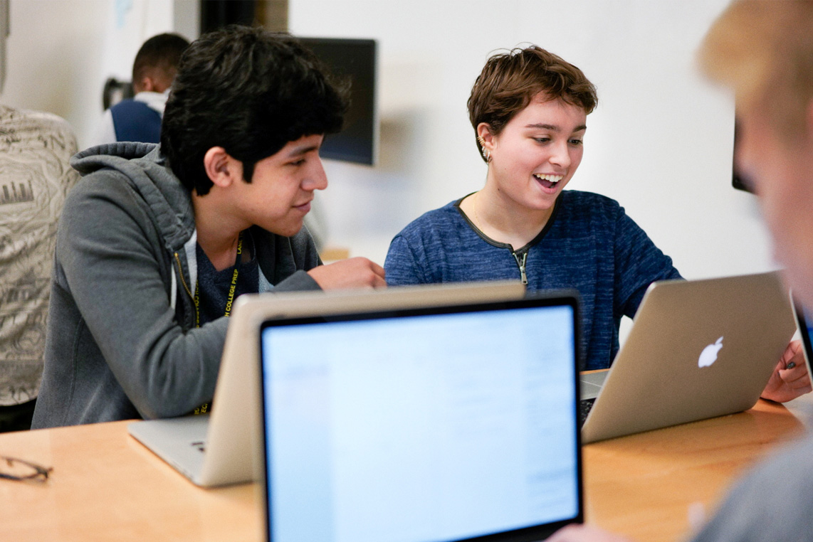Students in a classroom smiling looking at Apple laptops.