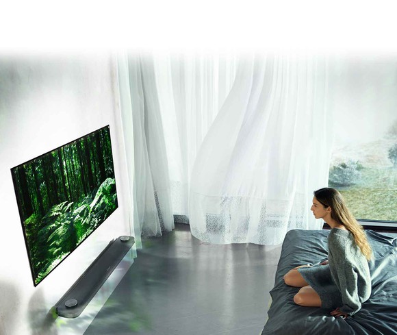 A woman sitting on a bed watching a 77" OLED TV from LG.
