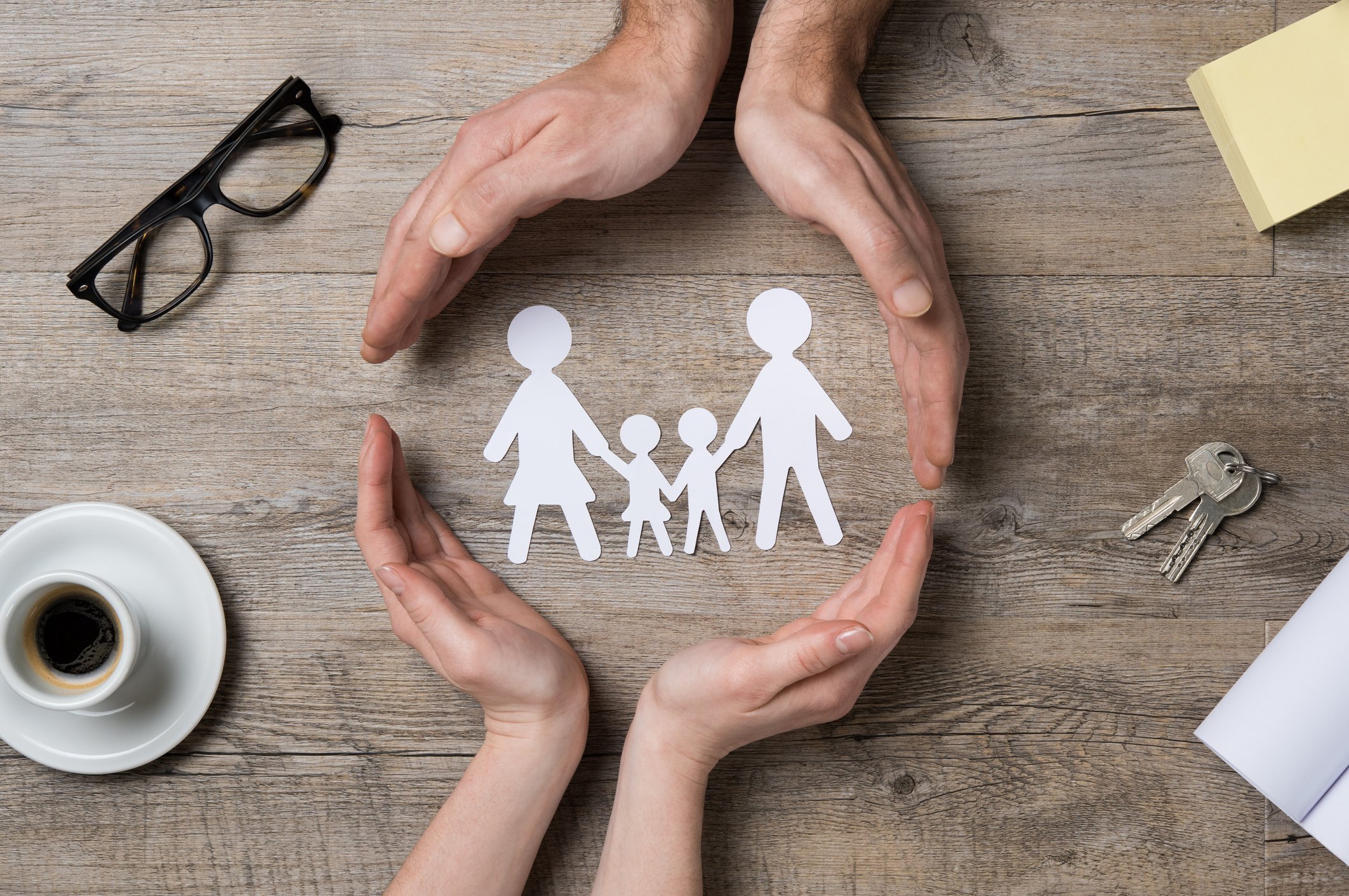 Two sets of hands making a circle around cut outs of paper figures of a man, woman, and two children holding hands.