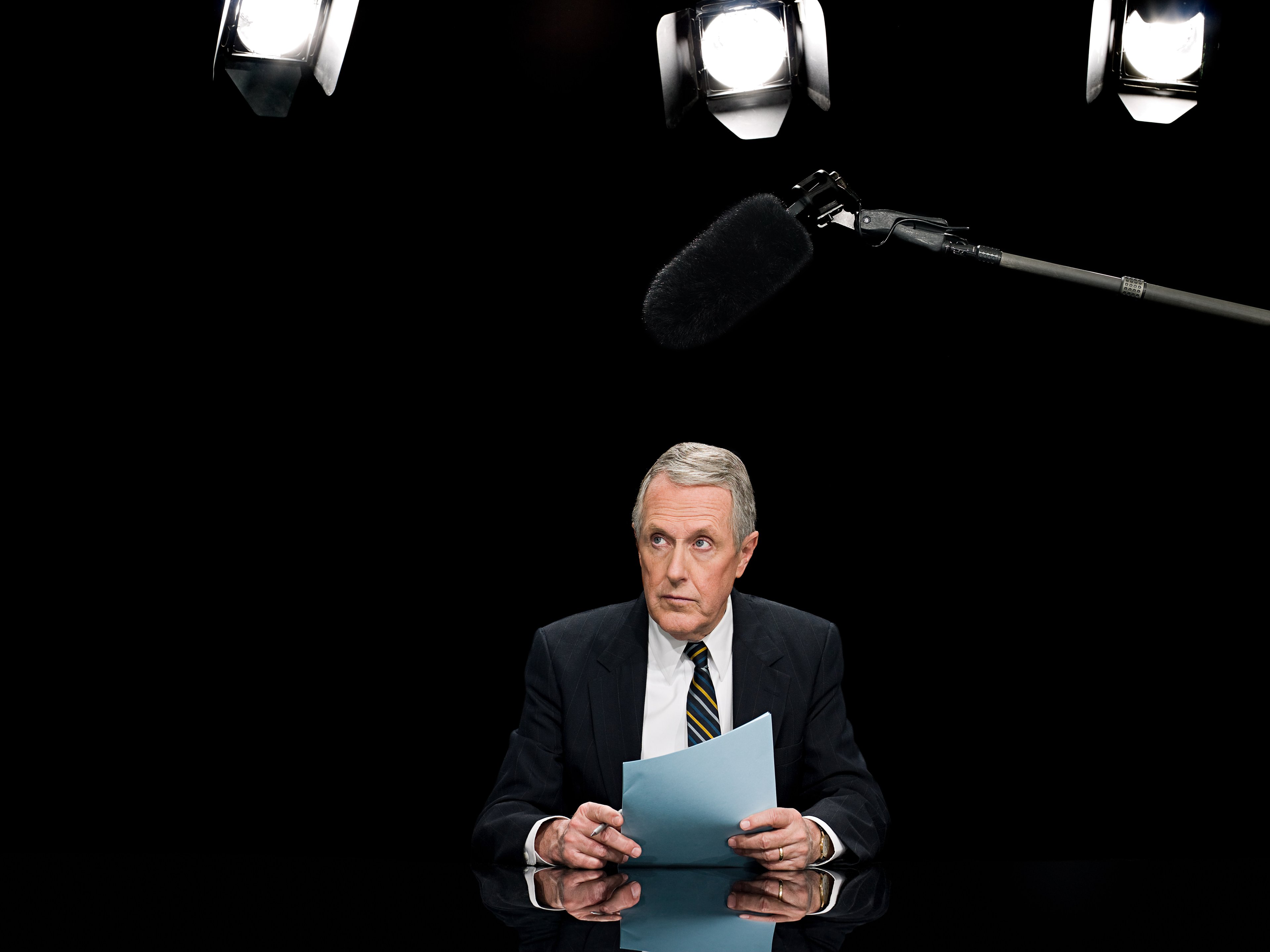 A man wearing a suit and holding a stack of papers is sitting behind a desk.