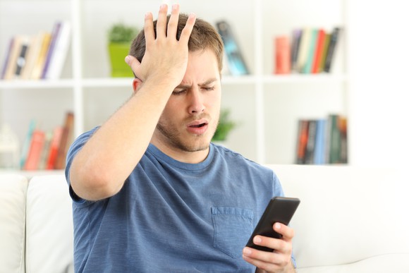 Young man holding a smartphone in one hand and face-palming with the other.