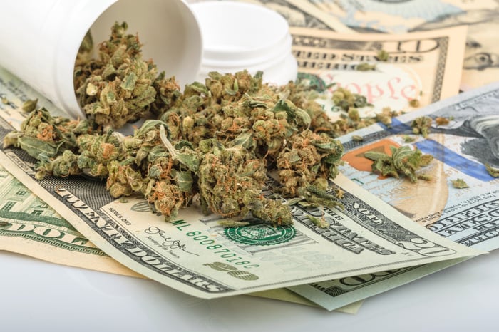 A bottle of dried cannabis buds tipped atop a messy pile of cash.