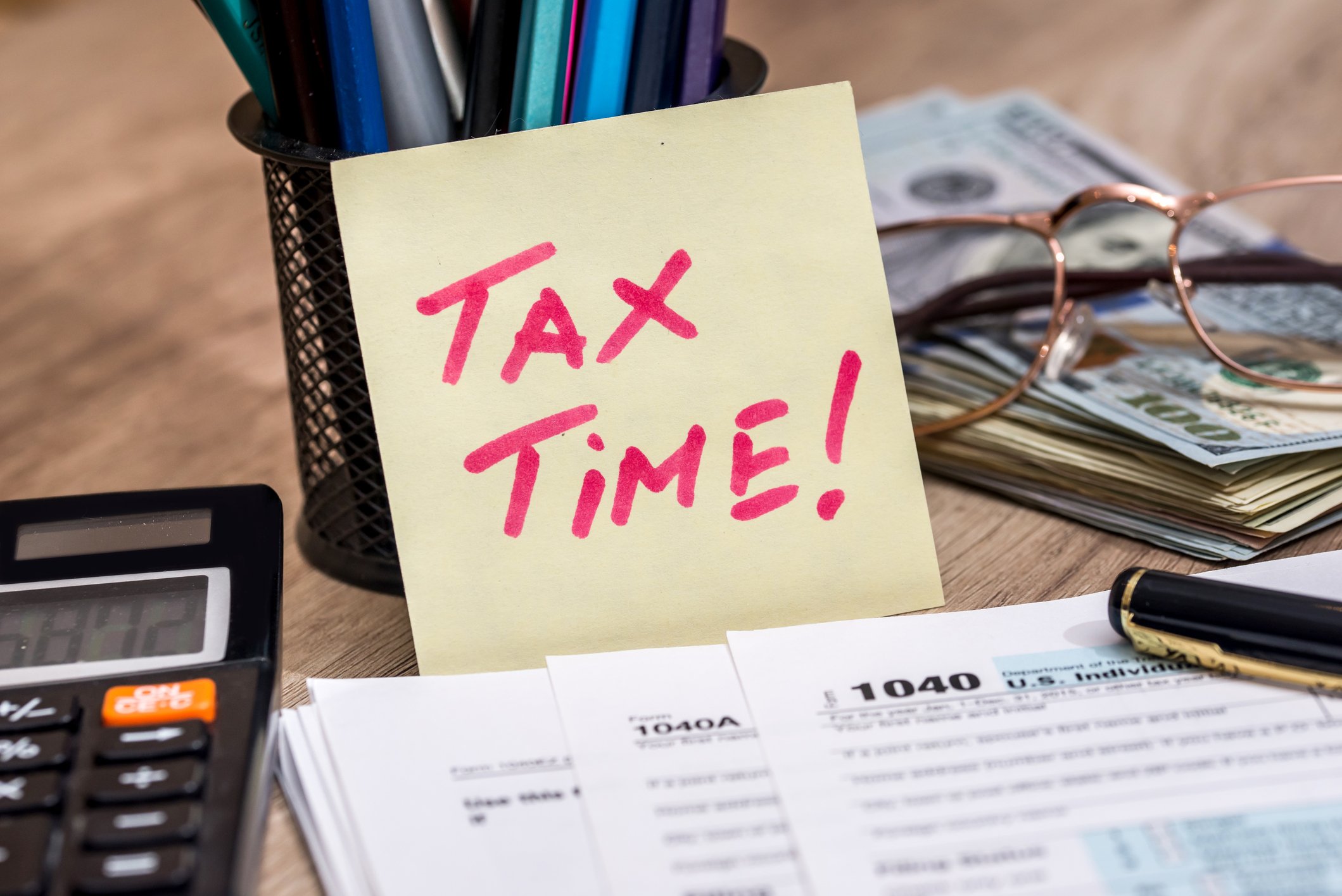 Post-it note reading tax time next to a can of pens, pile of money, calculator, and tax forms
