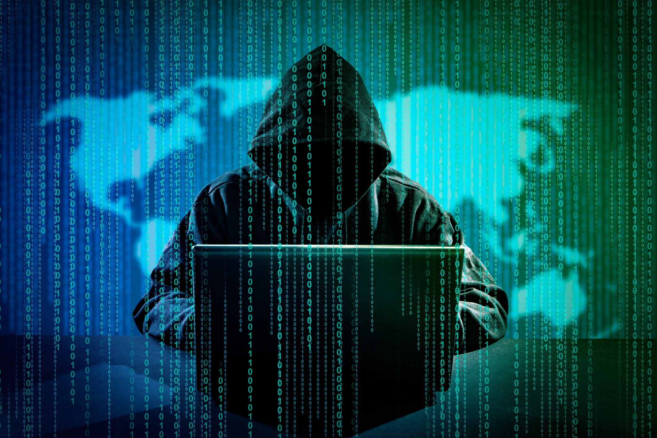 Hooded person sitting in front of computer with image of the world behind them.