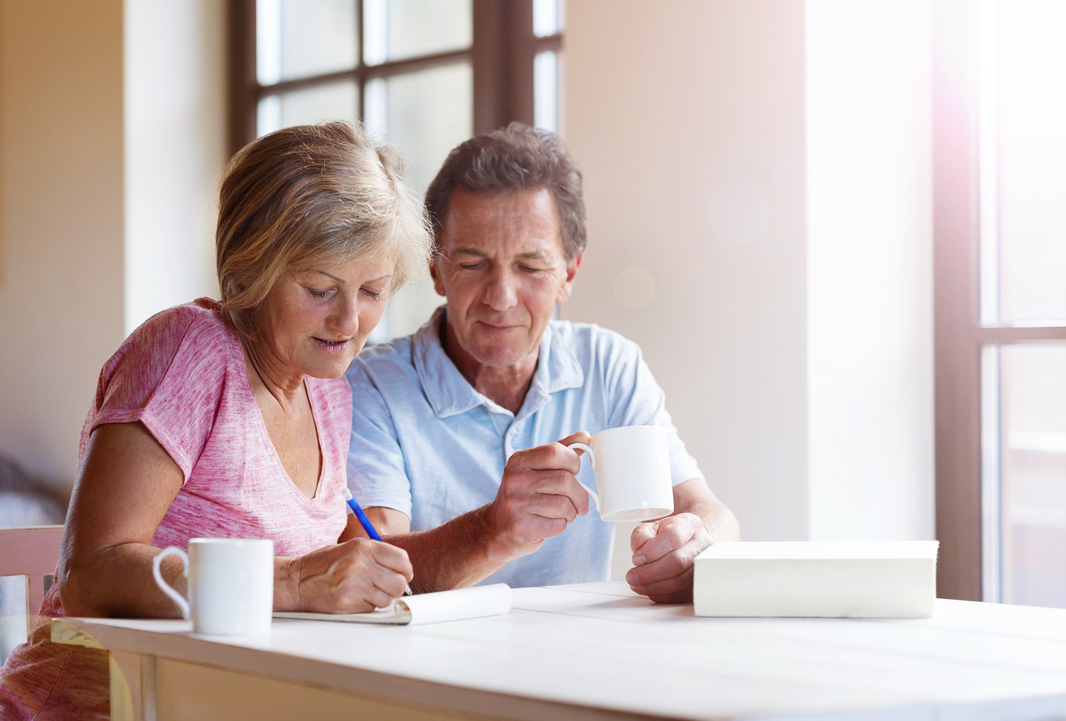 An older couple sits side by side near a window with light coming in. The man holds a coffee cup and looks on while the woman writes on a pad of paper.