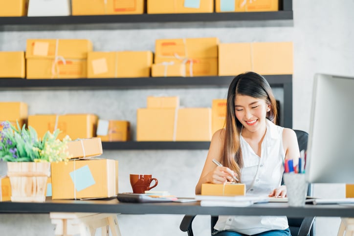 A young person seated at a desk, with boxes waiting to be shipped on shelving in background.