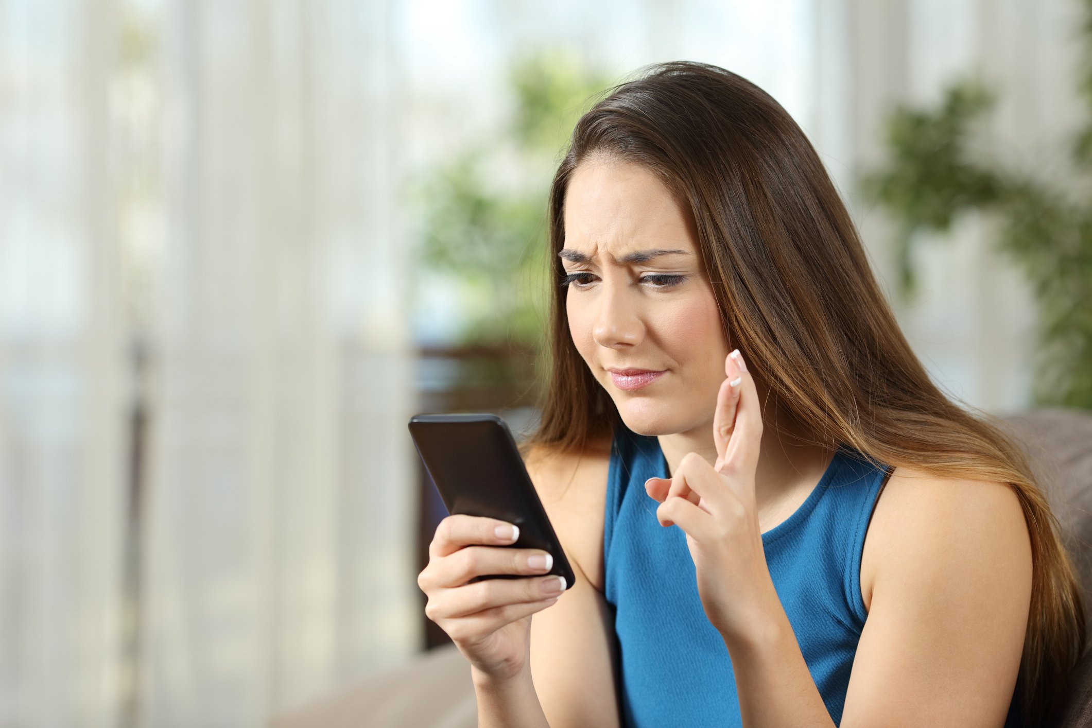 YOung woman frownning at her smartphone, fingers crossed.