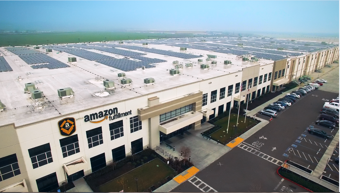 A huge Amazon fulfillment center complex showing bay doors for trucks, with a blue sky in background.