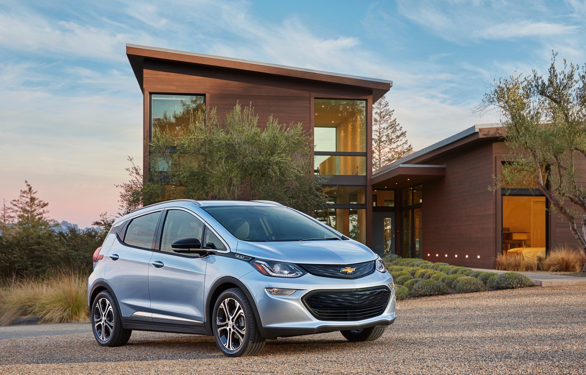 A light blue Chevrolet Bolt EV, a small electric hatchback, in front of an upscale house.