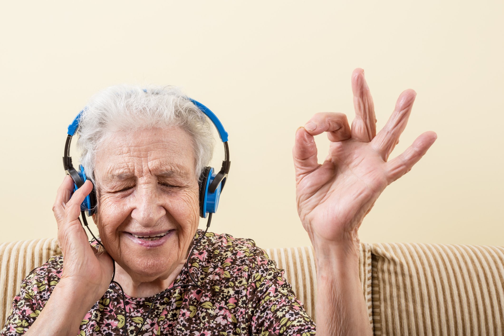 An elderly woman wearing headphones smiles as she makes the OK sign with her fingers.