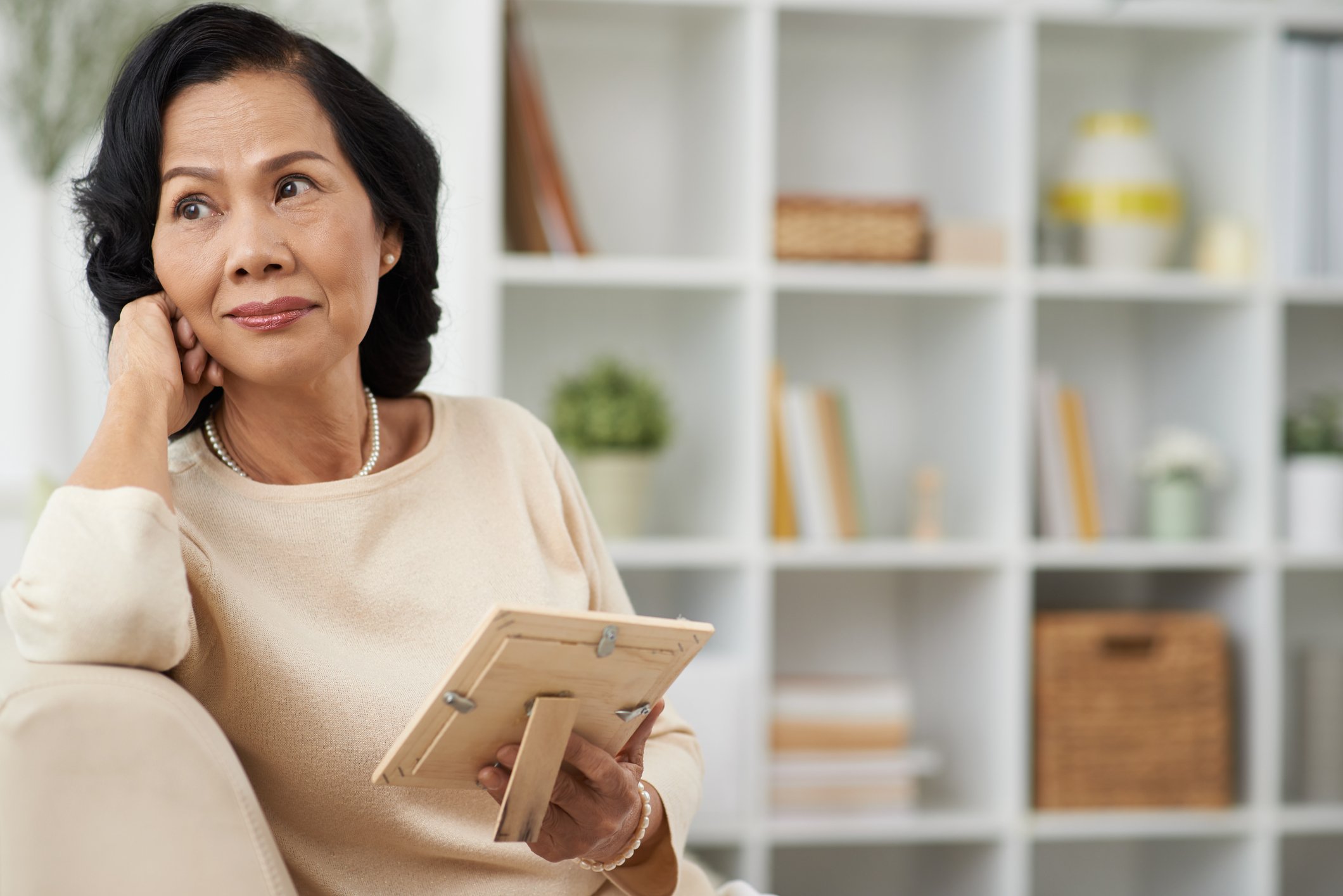 A mature woman holds a picture while sitting on a couch and looking off into the distance.