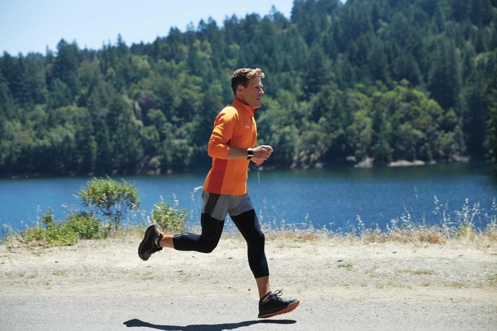 Man wearing a Fitbit and running along a lake with trees in the background.