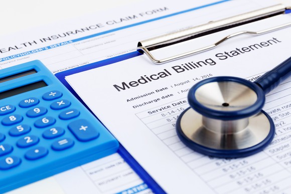 A Medical Billing Statement on a blue clipboard, next to a blue calculator, with a blue stethoscope sitting on top of it.