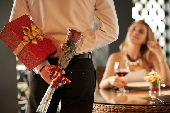 Man approaching a woman sitting at a table holding a rose and gift box behind his back