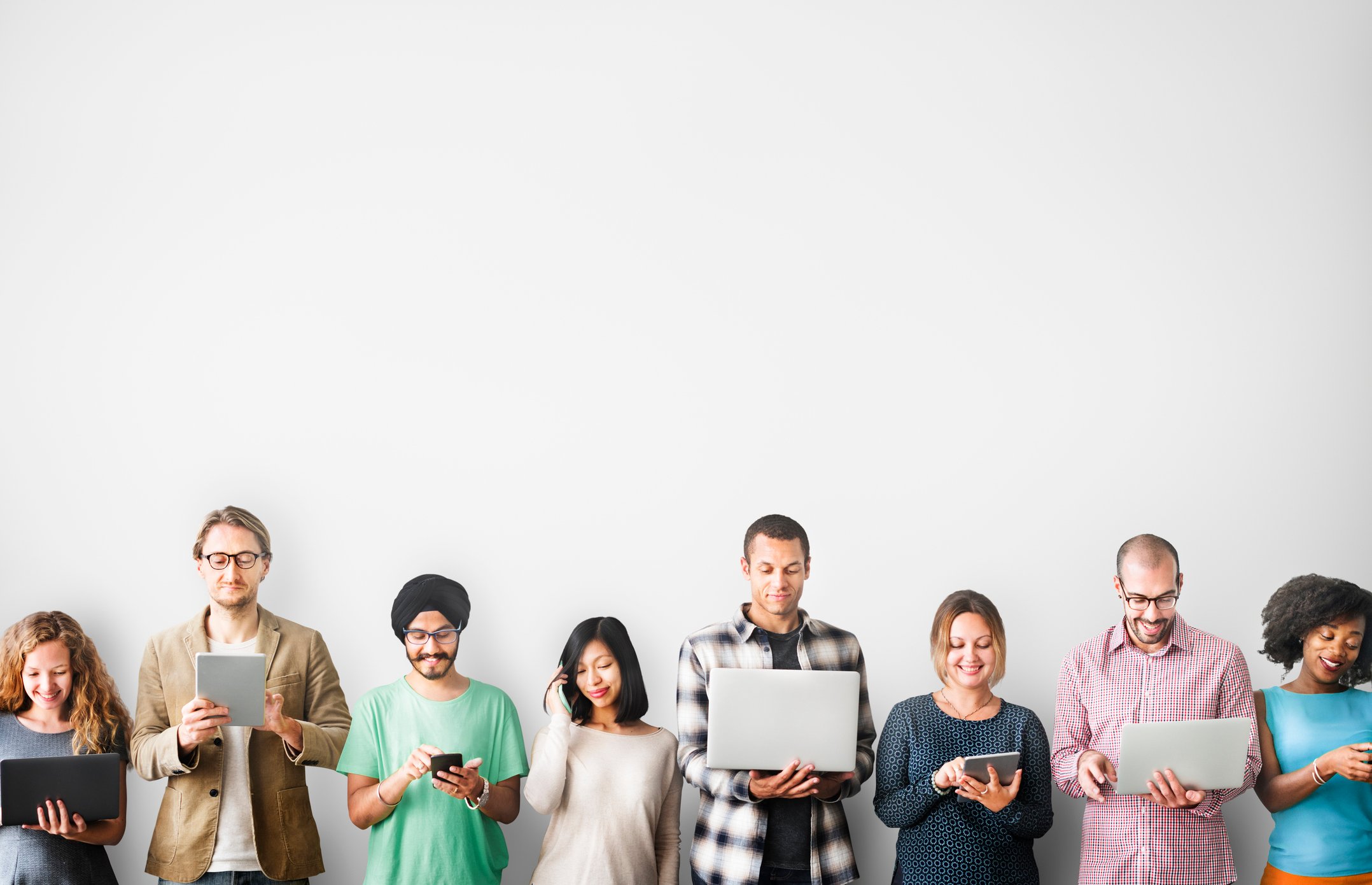 A group of people standing against a white wall using smartphones, tablets, and laptops.