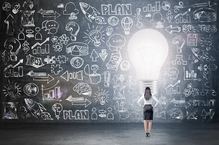 A woman reviewing a plan drawn on a massive chalkboard, surrounding a giant lightbulb