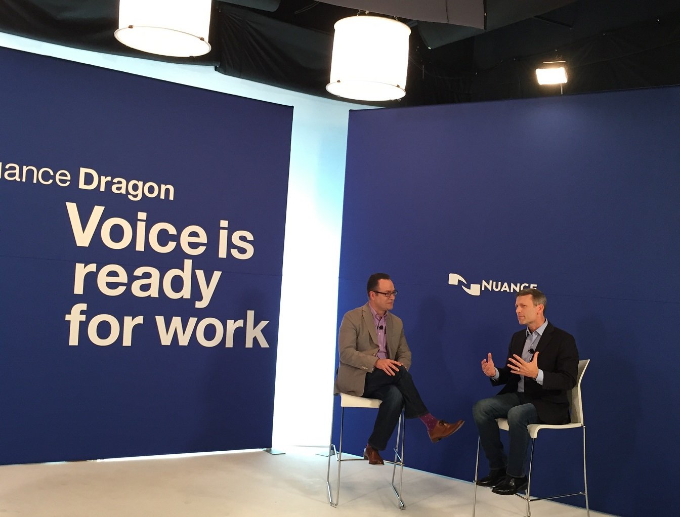 Two people in chairs in front of Nuance logo with slogan regarding Dragon on opposite blue wall.