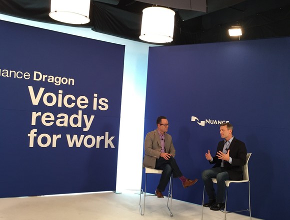 Two people in chairs in front of Nuance logo with slogan regarding Dragon on opposite blue wall.