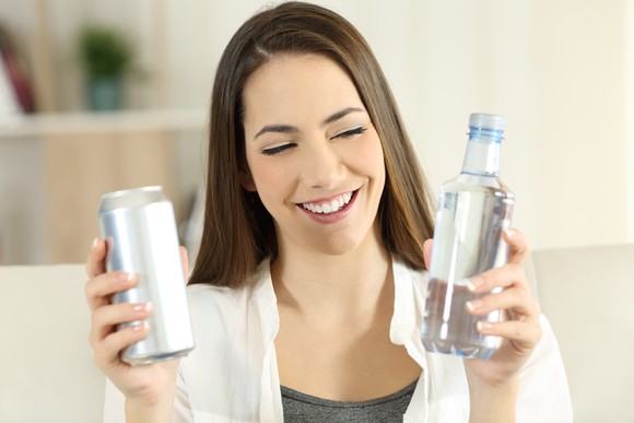 A smiling young woman chooses between water in a can or a bottle, both unbranded.