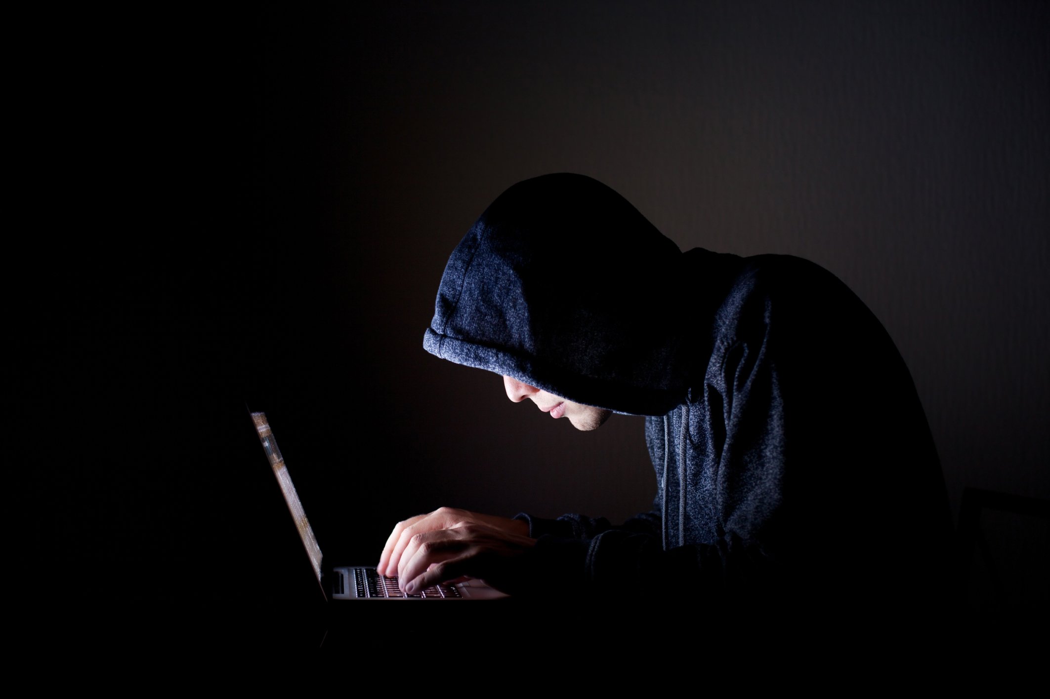 A hacker in a dark room hunched over a laptop with the glow of the screen highlighting his fingers and dark hoodie.