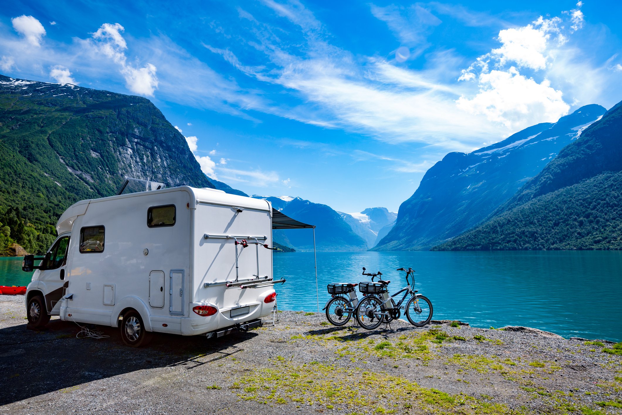 A white motorhome, with two bicycles next to it, parked next to a blue-green body of water and mountains in the background. 