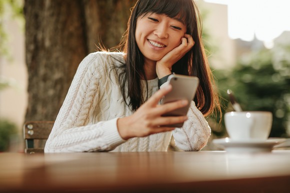 Chinese girl sitting at a table with a teacup in front of her, smiling at a smartphone
