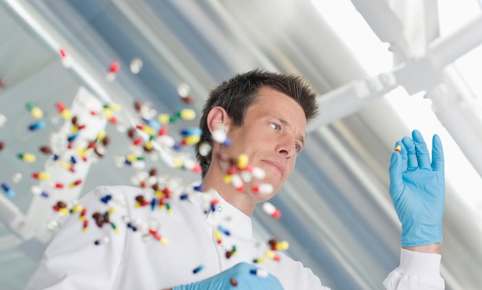 A man wearing a lab coat and blue disposable gloves sorting colorful pills on a glass table.