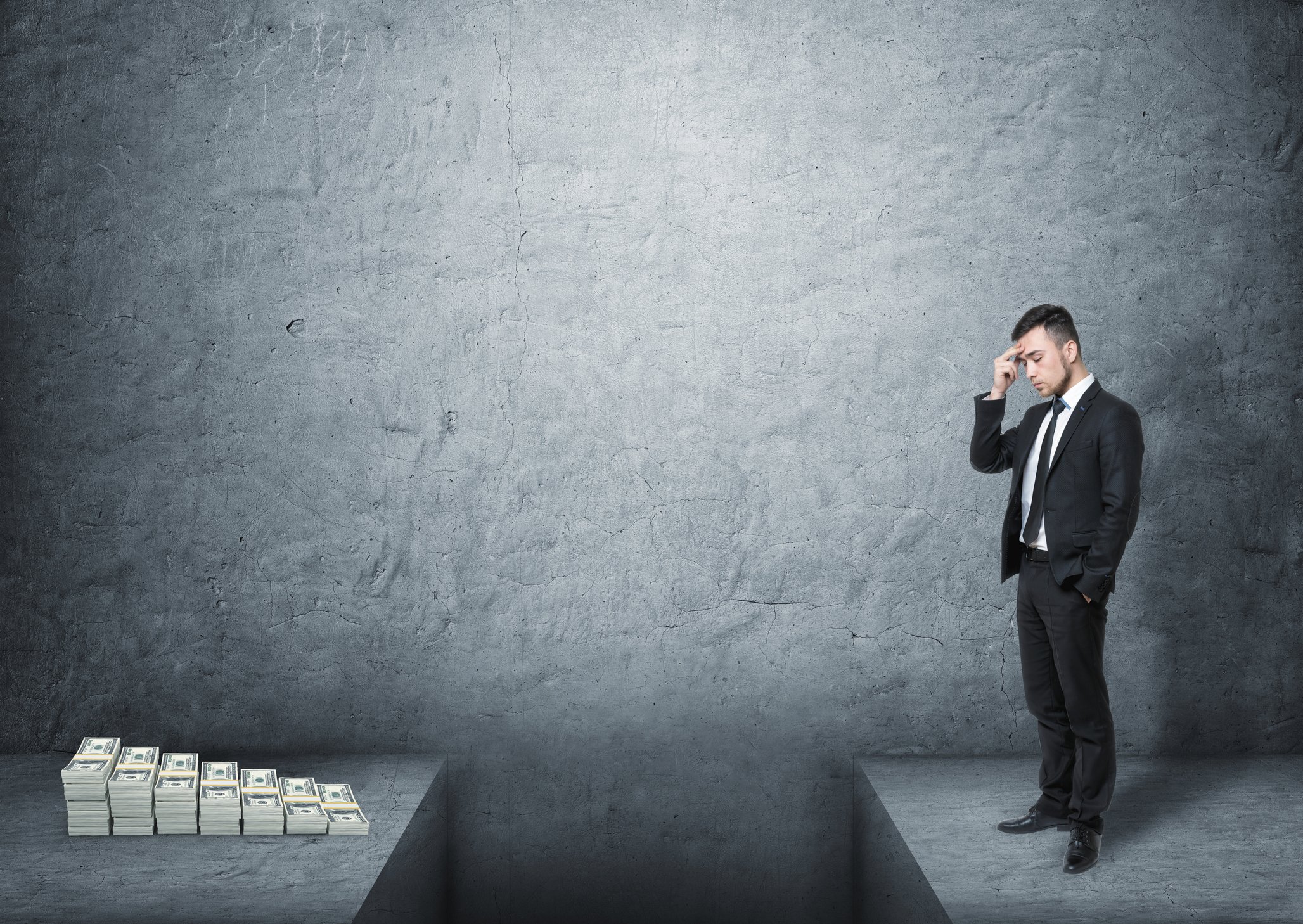 A frustrated man wearing a suit standing in front of a pile of money with a deep gap in the ground dividing them