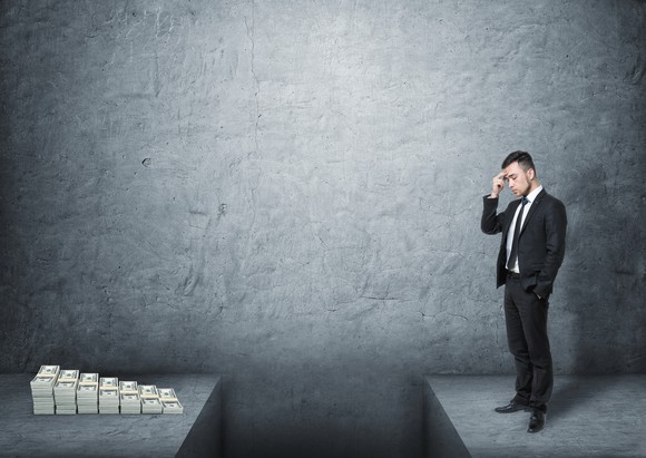 A frustrated man wearing a suit standing in front of a pile of money with a deep gap in the ground dividing them