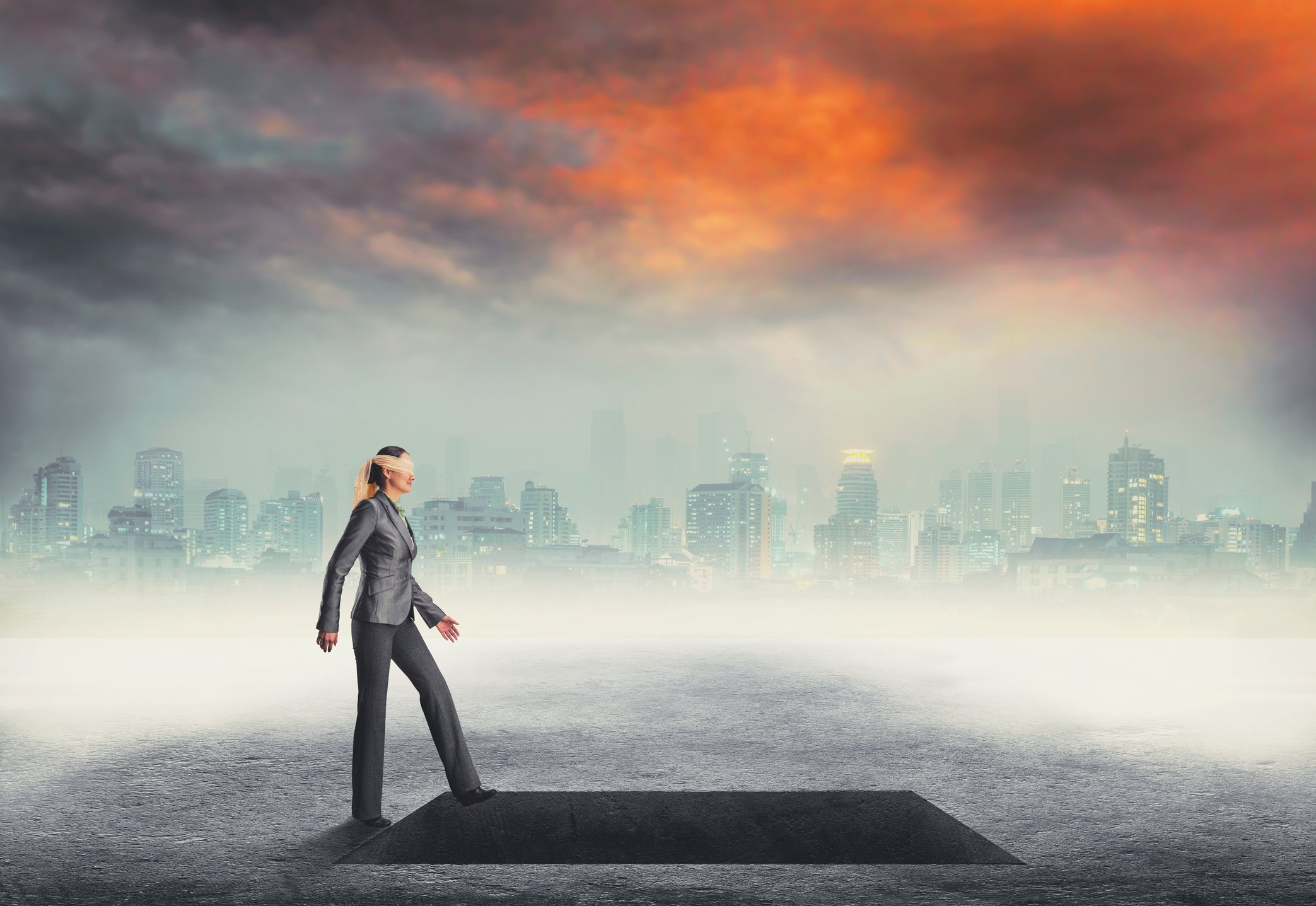 A blindfolded woman walking into a pit, with a city skyline at sunset in the background.