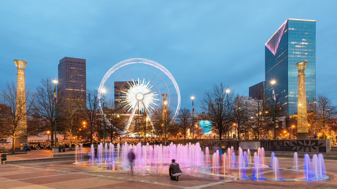 Purple lighted small fountains in front of a Ferris Wheel in a city park, with high buildings nearby.