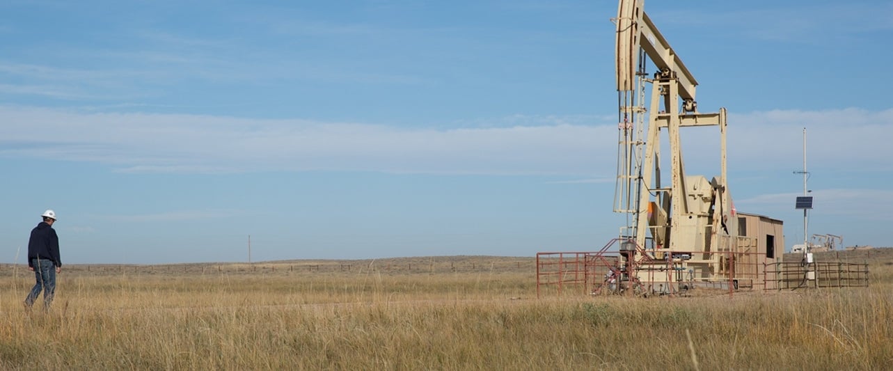 Well in grass field on clear day with worker wearing hard hat walking toward it.