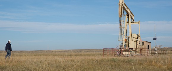 Well in grass field on clear day with worker wearing hard hat walking toward it.