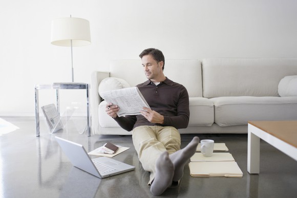 A man reading a newspaper while seated against a beige couch on his living room floor with an open laptop.