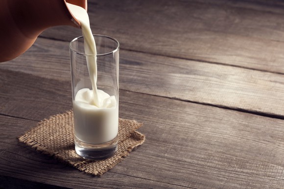 Pitcher pouring milk into a glass on a wood table with a cloth coaster under the glass.