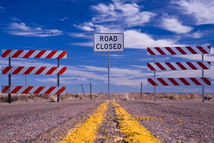 A road closed sign between two barricades on a rural road.
