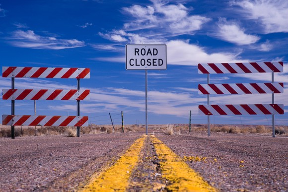 A road closed sign between two barricades on a rural road.