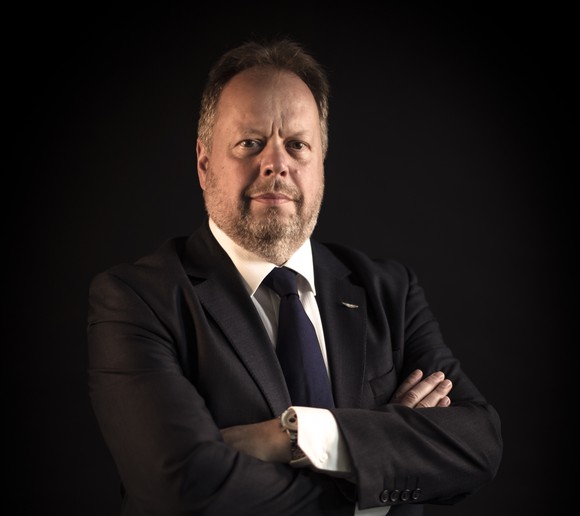 Palmer is standing, arms crossed, before a dark backdrop.