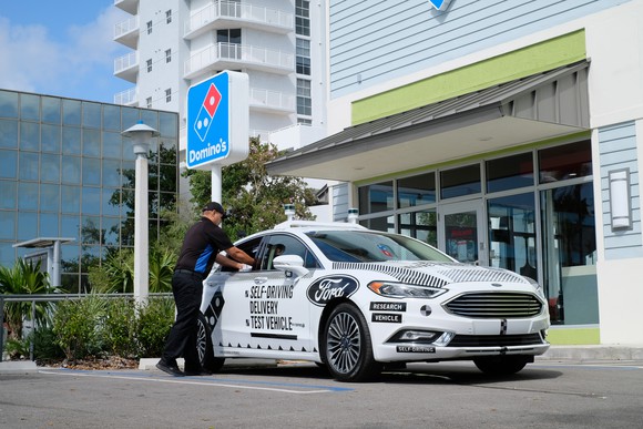 A white Ford Fusion with visible self-driving hardware is parked outside a Domino's Pizza restaurant. A Domino's employee is loading something into the vehicle via its passenger-seat window. 