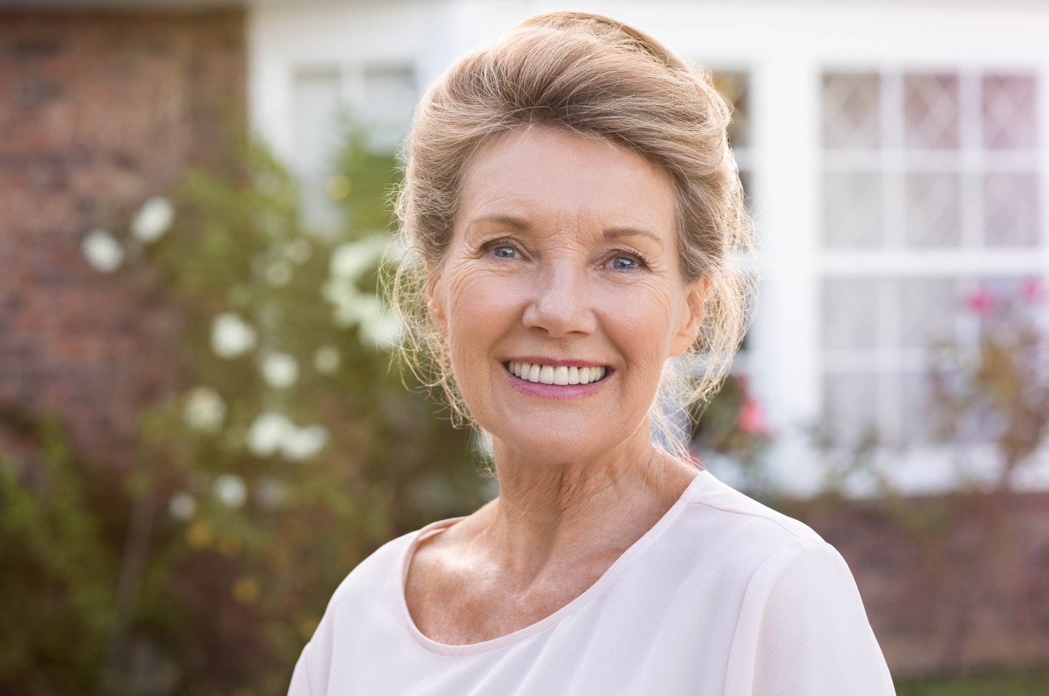 Smiling senior woman standing outdoors, with the windows of a house in the background.