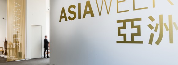 White wall with Asia Week in gold lettering, with a few exhibits behind a veil.