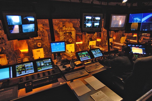 Control room with monitoring equipment above a strip steel mill manufacturing floor.